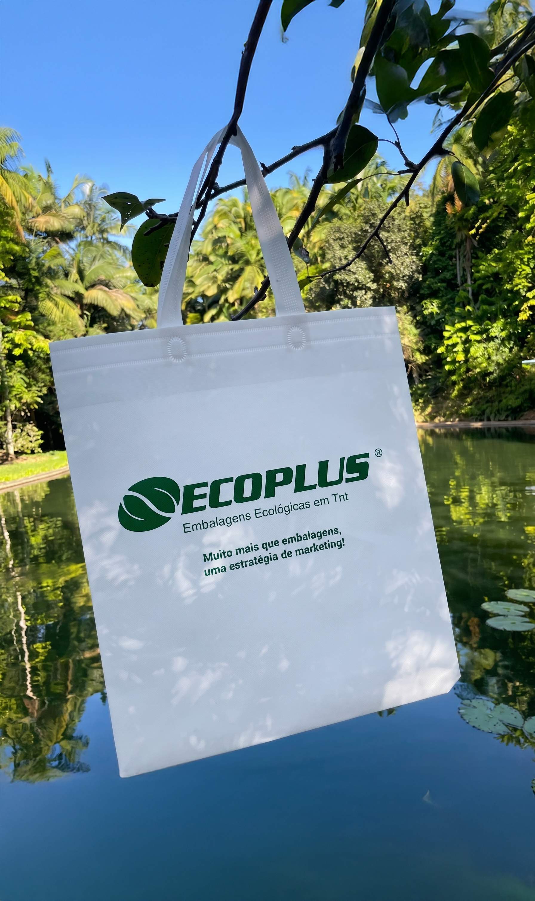 Sacola tote branca Ecoplus pendurada em galho sobre água com vegetação natural ao fundo